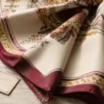 Vintage silk scarf folded and partially opened on a neutral surface, showing printed border, rolled hem, color details and textile texture.
